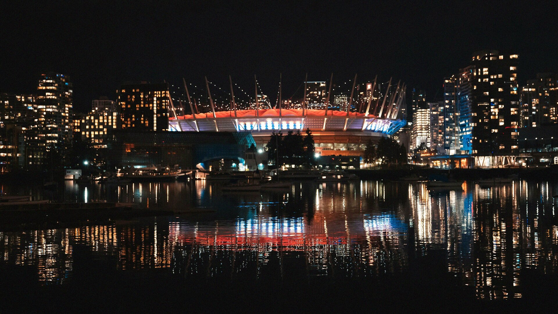 BC Place stadium glowing red and white at night with the downtown Vancouver skyline reflected in False Creek
