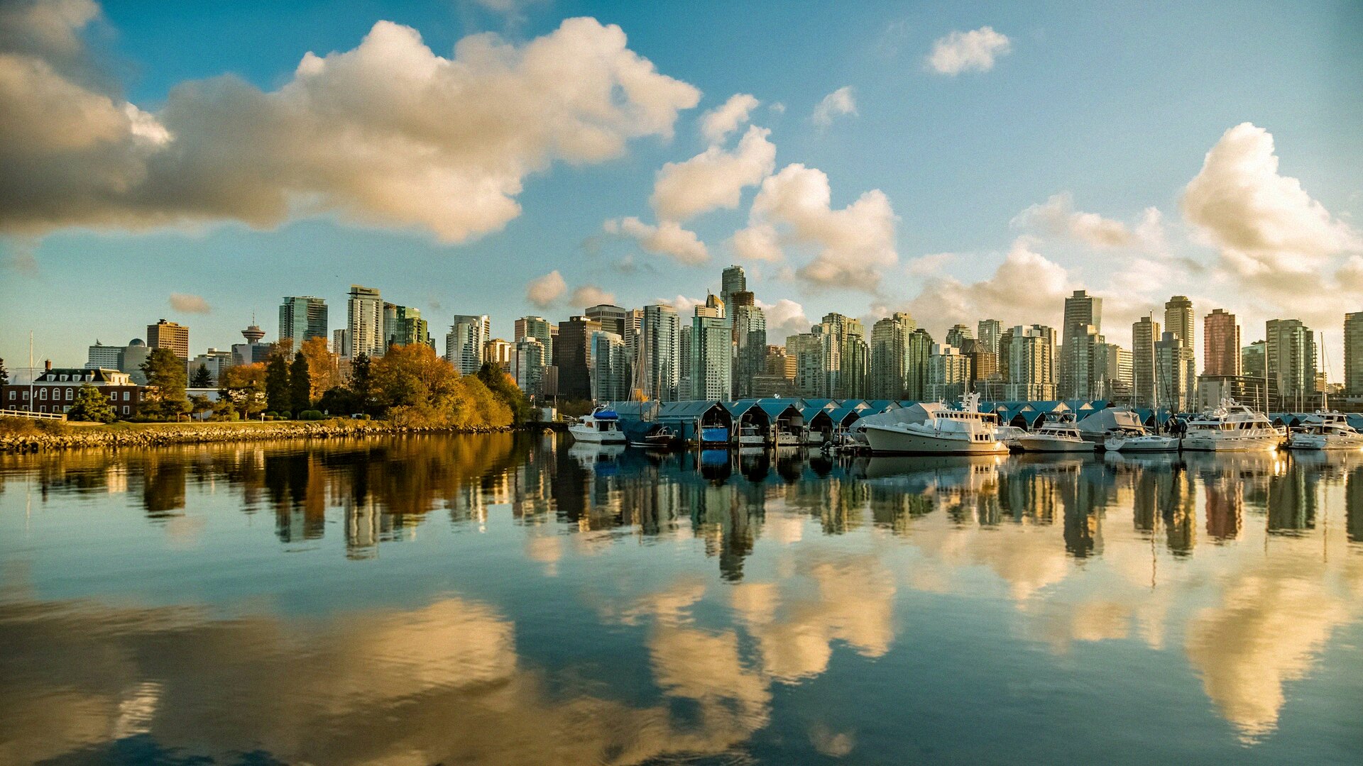 The downtown Vancouver skyline at golden hour reflected in calm Coal Harbour water with sailboats moored along the seawall