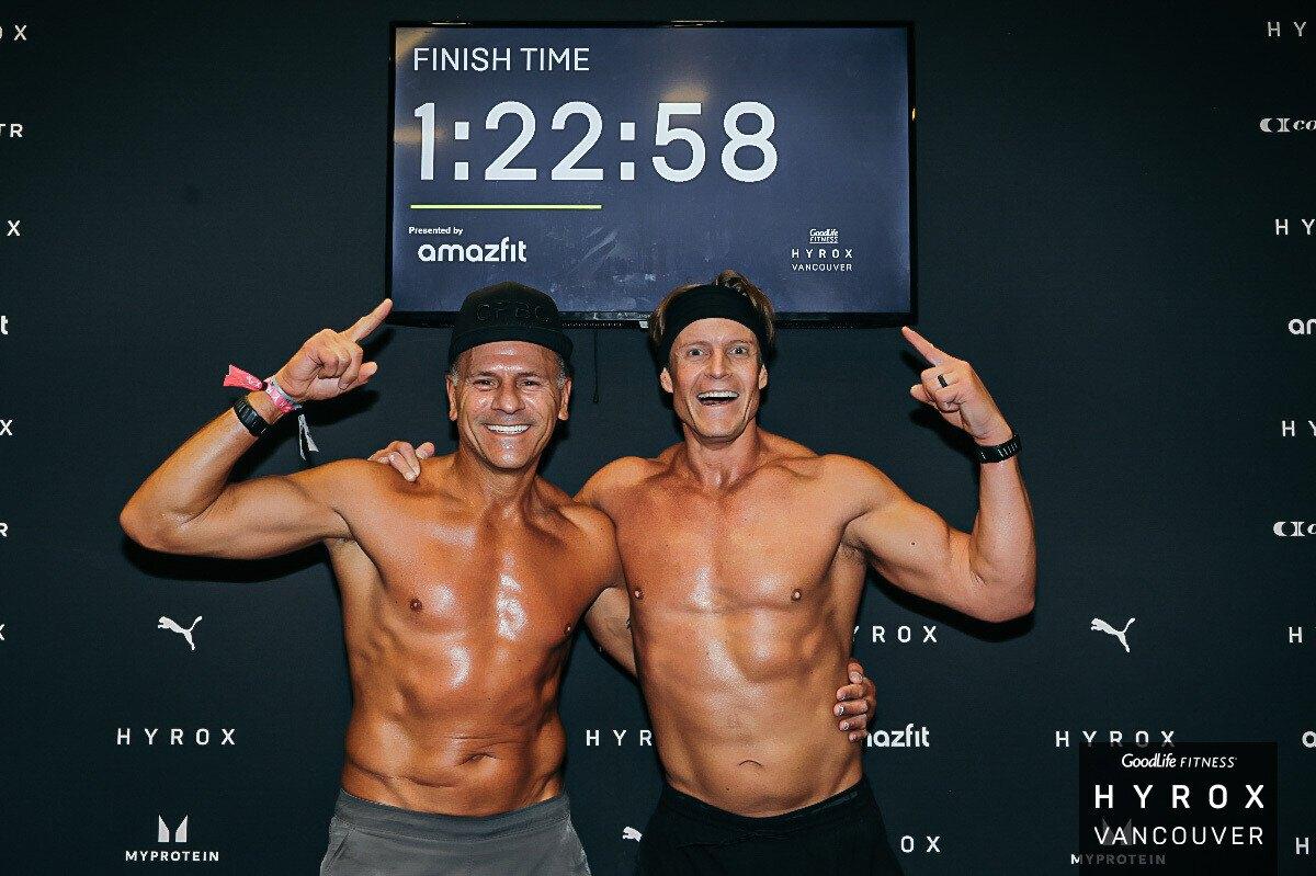 Two CrossFit BC athletes celebrating in front of the finish time board at HYROX Vancouver